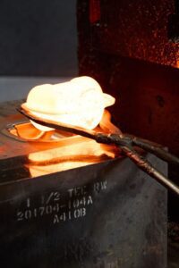 A pair of tongs holds a glowing hot metal object above a mold, showing the process of metal forging or casting, with intense orange light from the heated materials.