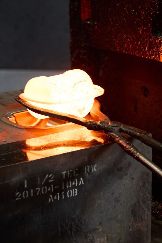 A pair of tongs holds a glowing hot metal object above a mold, showing the process of metal forging or casting, with intense orange light from the heated materials.