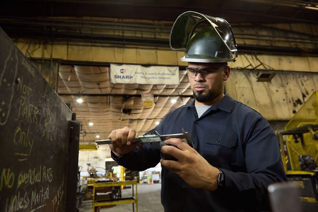 A worker in safety gear and glasses uses a caliper to measure an object in an industrial warehouse, with equipment and a SHARP safety banner visible in the background.