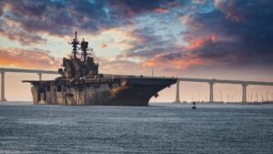 A large naval ship is anchored in a bay at sunset, with dramatic clouds in the sky and a long bridge in the background. Sailboats are docked near the shore, highlighting the area’s role in maritime defense.
