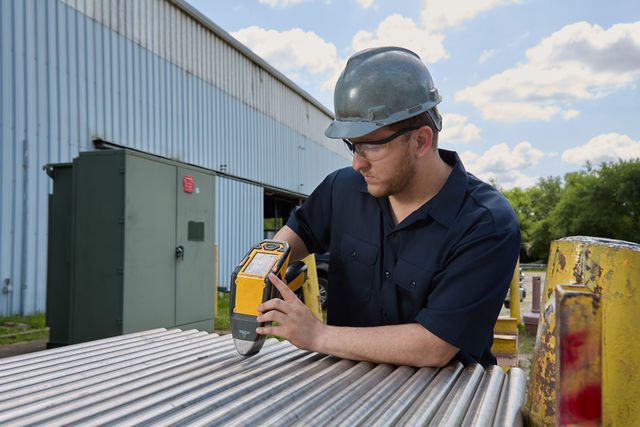 A man wearing safety glasses, a hard hat, and a navy shirt uses a handheld scanning device on metal pipes outdoors near a large industrial building.