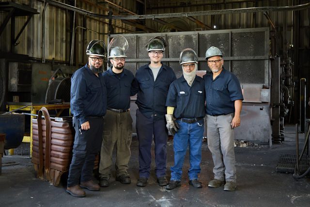 Five industrial workers in protective gear and helmets stand together, smiling, inside a factory with metal equipment and machinery in the background.