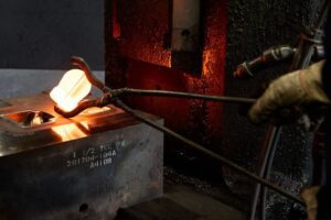 A gloved hand uses tongs to hold a glowing hot metal piece over a mold in an industrial forging setting, with molten metal, machinery, and elements of power generation visible.