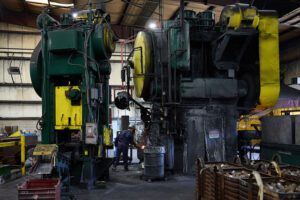 A worker at U.S. Drop Forge operates large press hammer in a factory setting, surrounded by metal equipment and tools.